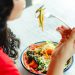 A woman in a red shirt enjoys a fresh salad indoors at a restaurant table.