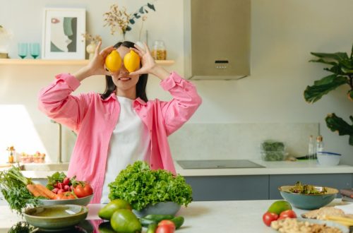 A woman playfully holding lemons over her eyes in a bright, lively kitchen setting filled with fresh vegetables.